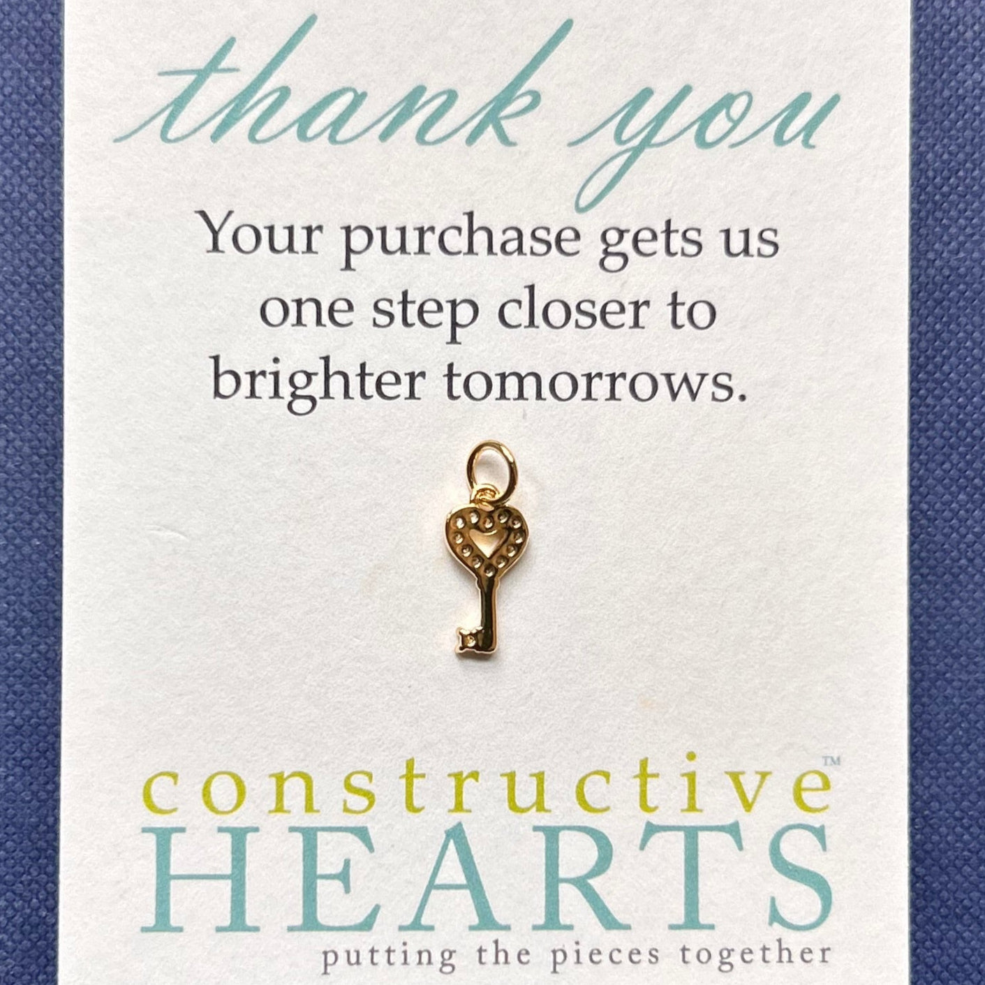 Gold key-shaped pave charm on a card with 'constructive HEARTS' branding and a message.